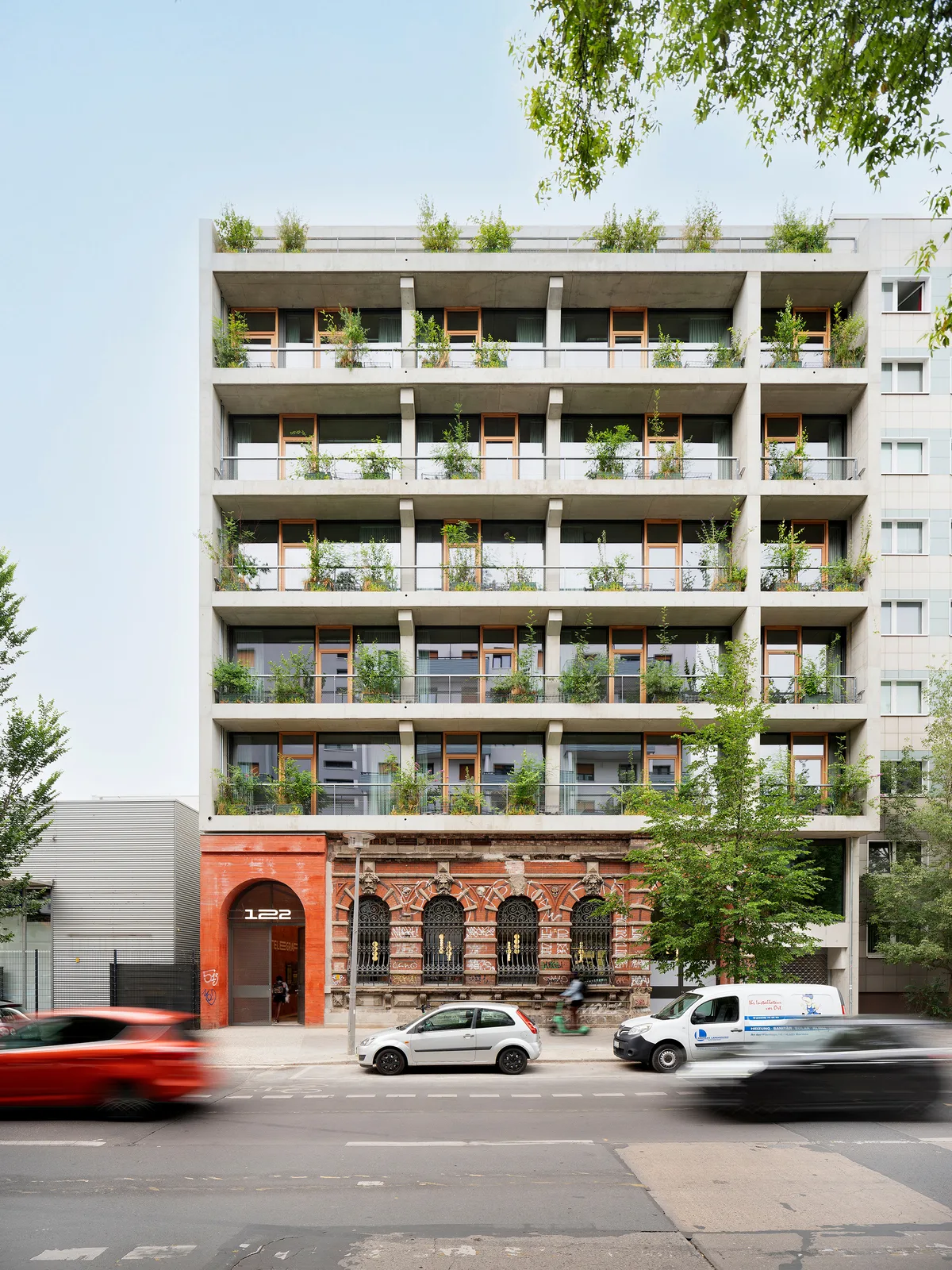Moderne Architektur trifft auf historische Bausubstanz: Ein nachhaltiges Wohngebäude mit begrünten Balkonen erhebt sich über einer restaurierten Backsteinfassade. Der Kontrast zwischen dem minimalistischen Neubau mit großen Glasfronten und dem denkmalgeschützten Sockel aus rotem Ziegelstein zeigt die gelungene Verbindung von Alt und Neu in der urbanen Architektur.