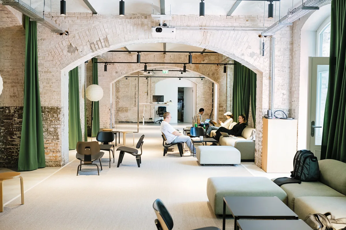 Modernes Coworking-Space in einem Loft mit Backsteinwänden, grünen Vorhängen und stilvollen Möbeln. Mehrere Personen arbeiten und unterhalten sich in einer offenen, kreativen Umgebung mit bequemen Sitzgelegenheiten, Tischen und Laptops.