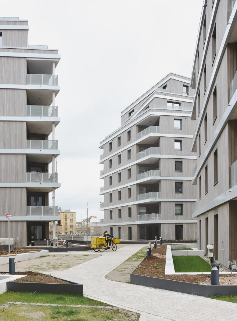 Innenhof des Wohnquartiers mit mehrgeschossigen Holzfassaden, auskragenden Balkonen mit Streckmetall-Brüstungen, gepflasterten Wegen und Pflanzbeeten – Wohnungsbau im Quartier.