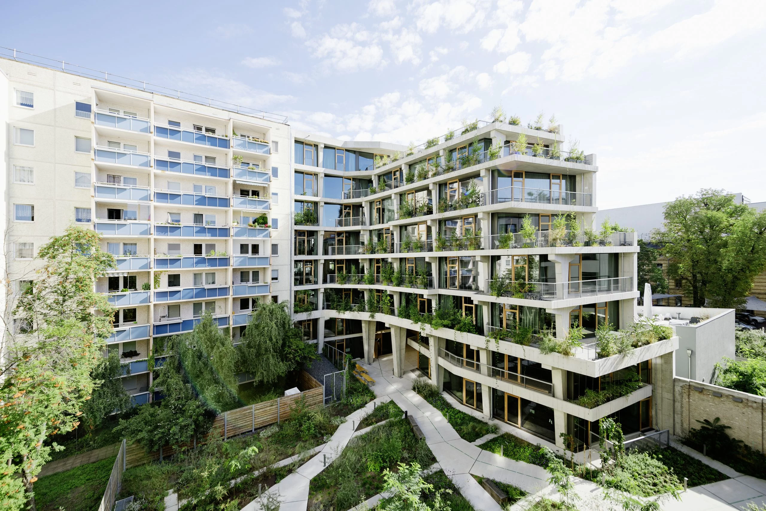 Nachhaltige Architektur im urbanen Raum: Ein modernes Wohngebäude mit terrassenförmiger Struktur und üppiger Begrünung integriert sich harmonisch in die Umgebung. Große Glasfronten und Holzfenster sorgen für lichtdurchflutete Innenräume, während die bepflanzten Balkone das Konzept des vertikalen Gartens betonen. Ein begrünter Innenhof mit organisch verlaufenden Wegen fördert das nachhaltige Wohnen und schafft eine naturnahe Atmosphäre in der Stadt.