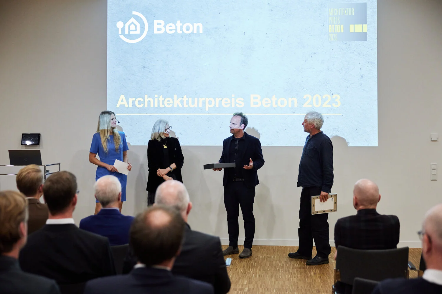 Verleihung des Architekturpreises Beton 2023 – Preisträger stehen auf der Bühne und nehmen die Auszeichnung entgegen. Vor einer Leinwand mit dem Logo und Titel der Veranstaltung diskutieren die Gewinner mit den Moderatorinnen über ihr innovatives Architekturprojekt. Der Architekturpreis Beton würdigt herausragende Bauwerke, die mit nachhaltiger und kreativer Betonbauweise überzeugen. Die Veranstaltung zieht zahlreiche Fachleute aus Architektur und Bauwesen an.