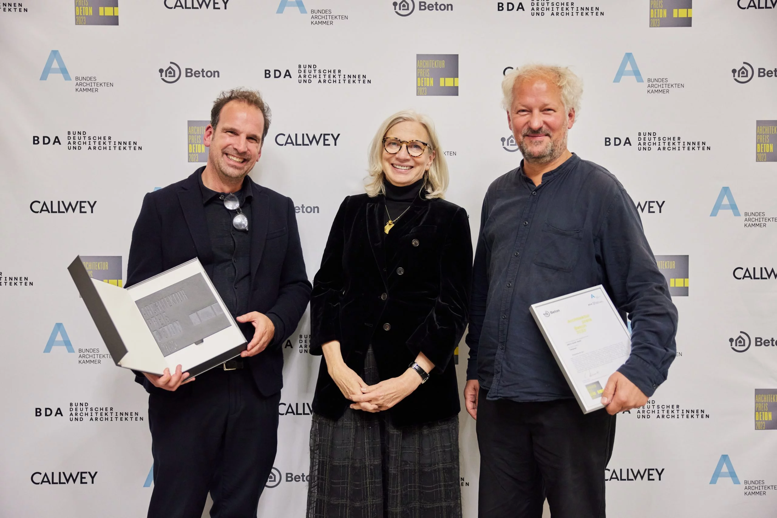 Preisverleihung des Architekturpreises Beton 2023 mit zwei strahlenden Preisträgern und der Vorsitzenden der Jury vor einer Sponsorenwand. Die ausgezeichneten Architekten halten ihre Urkunden und die Gewinnerplakette in den Händen. Die Veranstaltung würdigt herausragende Architekturprojekte, die innovative und nachhaltige Betonbauweise einsetzen. Organisiert vom Bund Deutscher Architektinnen und Architekten (BDA) sowie der Bundesarchitektenkammer, zählt der Preis zu den renommiertesten Auszeichnungen in der Baubranche.