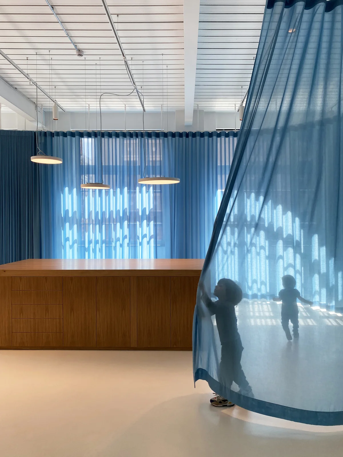 Modernes Interieurdesign mit lichtdurchflutetem Raum und bodentiefen, transparenten blauen Vorhängen. Die offene Architektur kombiniert minimalistische Ästhetik mit funktionaler Raumgestaltung. Eine elegante Holztheke setzt einen warmen Kontrast zur klaren, industriellen Decke mit sichtbaren Metallstrukturen und hängenden Designerleuchten. Die spielenden Kinder hinter dem Vorhang erzeugen eine atmosphärische Licht-Schatten-Interaktion, die die Transparenz und Leichtigkeit des Raumkonzepts betont. Perfekte Verbindung aus skandinavischem Design, flexibler Raumaufteilung und hochwertiger Materialwahl für moderne Büro- oder Wohnkonzepte.