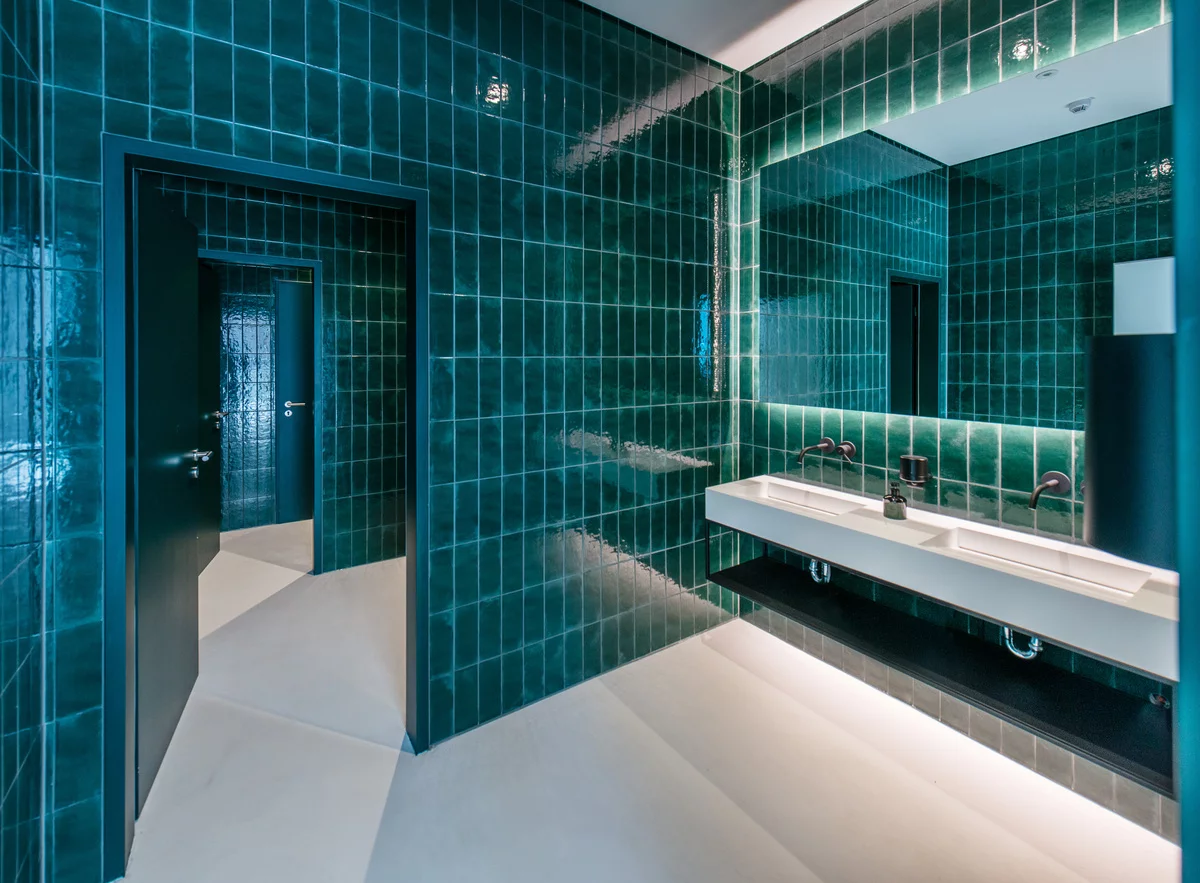 Modern gestaltetes Badezimmer mit elegantem Architekturdesign. Die tiefgrünen, glänzenden Wandfliesen verleihen dem Raum eine luxuriöse Atmosphäre und setzen stilvolle Akzente. Ein minimalistisches Doppelwaschbecken mit integrierter LED-Beleuchtung sorgt für eine moderne Ästhetik und eine angenehme Lichtstimmung. Die Kombination aus dunklen Türen und kontrastreichen, hellen Bodenflächen schafft ein harmonisches Raumgefühl. Die cleane Linienführung und die hochwertige Materialwahl unterstreichen den exklusiven Charakter dieses Sanitärbereichs. Ideal für moderne Büro- oder Hotelarchitektur mit einem Fokus auf Design und Funktionalität.