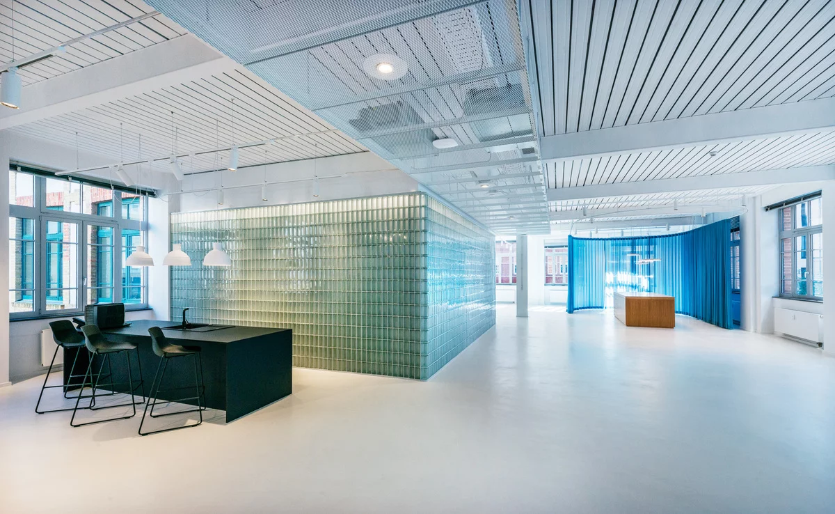 Modernes Loft-Büro im LUX Tower Berlin mit innovativer Architektur und industriellem Charme. Die offene Raumgestaltung kombiniert gläserne Bauelemente mit minimalistischer Ästhetik. Eine transluzente Glasbausteinwand dient als stilvolles Raumtrenner-Element und sorgt für eine helle, lichtdurchflutete Atmosphäre. Die freihängenden weißen Pendelleuchten über der schwarzen Kücheninsel setzen moderne Akzente und betonen das kontrastreiche Design. Hohe Decken mit einer offenen Gitterstruktur verleihen dem Raum eine luftige Weite, während blaue Vorhänge flexible Rückzugsorte schaffen. Die großen Fenster mit Blick auf die historische Backsteinfassade verbinden Altbaucharme mit zeitgemäßer Büroarchitektur.