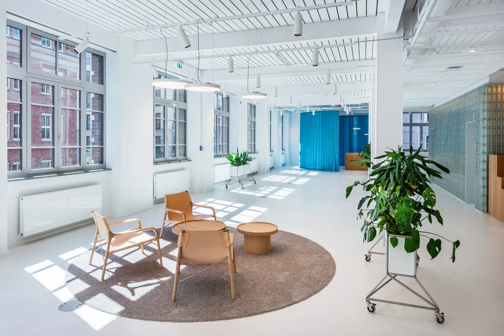 Modernes Bürodesign im LUX Tower Berlin mit lichtdurchflutetem, offenem Raumkonzept. Die hohe Decke mit sichtbarer Stahlkonstruktion und Hängeleuchten unterstreicht den industriellen Charme des historischen Gebäudes. Große Fenster mit grauen Metallrahmen lassen viel Tageslicht herein und schaffen eine helle, einladende Atmosphäre. Blaue Vorhänge setzen farbliche Akzente und bieten flexible Raumtrennung. Natürliche Materialien wie Holz und Leder ergänzen das minimalistische, skandinavisch inspirierte Interior. Die architektonische Verbindung von Alt und Neu macht dieses Büro zu einem modernen Arbeitsumfeld mit historischem Charakter.