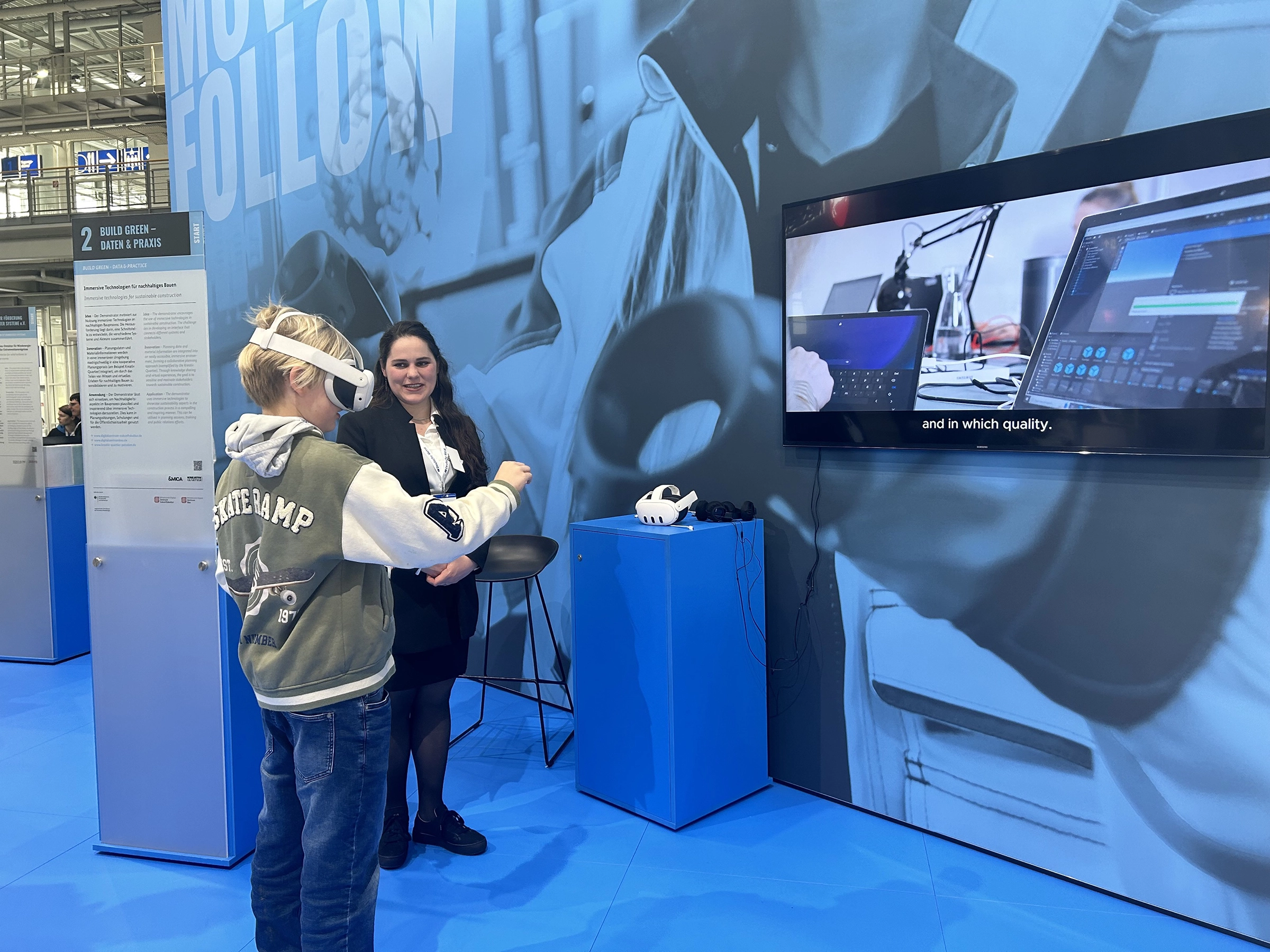 Virtuelle Realität auf der Messe: Ein Junge testet eine VR-Brille an einem interaktiven Messestand zum Thema digitale Technologien und nachhaltiges Bauen. Im Hintergrund erklärt eine Messebetreuerin das Konzept, während ein großer Bildschirm mit einer Software-Demonstration moderne Architektur- und Planungsprozesse veranschaulicht. Der Stand kombiniert innovative Technik mit interaktiven Erlebnissen.