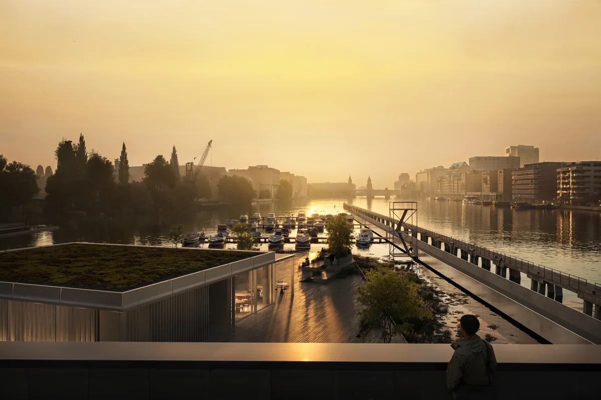 Stadtlandschaft bei Sonnenuntergang: Auf dem Bild ist ein moderner, langgestreckter Steg oder Gehweg zu sehen, der über Wasser führt. Rechts und links davon gibt es Begrünung und Sitzgelegenheiten. Im Hintergrund sind Gebäude, Bäume und ein Kran zu erkennen, alles in warmes, goldenes Sonnenlicht getaucht. Menschen sind auf dem Weg unterwegs, einige sitzen am Rand. Die Atmosphäre wirkt ruhig und stimmungsvoll.