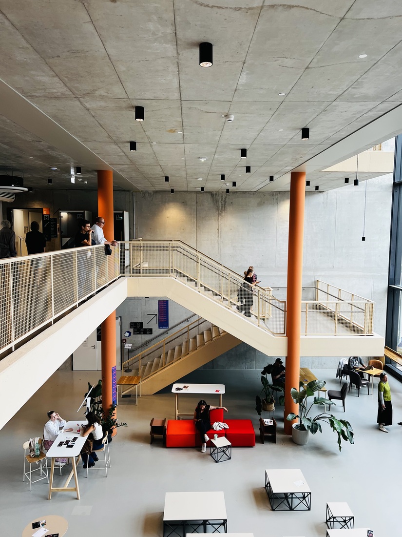 Offener Gemeinschaftsbereich im Kreativ Quartier Potsdam mit Sichtbetondecke, orangefarbenen Stützen, geschwungener Treppe und flexiblen Arbeitsplätzen.