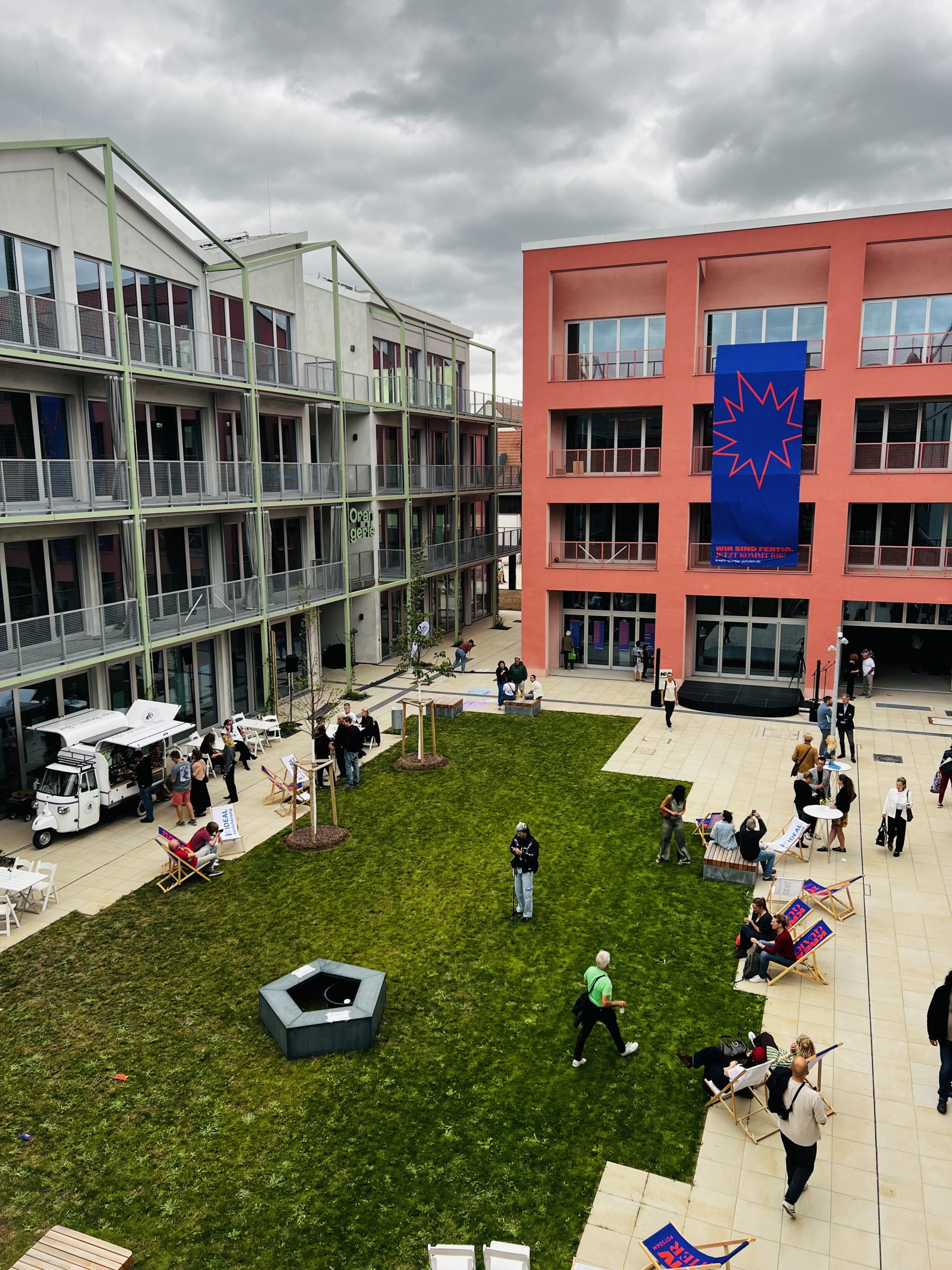 Eröffnungsfeier im Kreativ Quartier Potsdam: Blick von oben auf den zentralen Rasenplatz mit Gästen, Liegestühlen und dem Banner „Wir sind fertig" an der Fassade der Akademie.